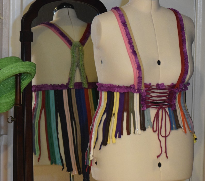 May include: A purple and white corset with a pink and white trim. The corset is made of a variety of colorful strips of fabric, possibly zippers. The corset is tied at the back with a burgundy ribbon.