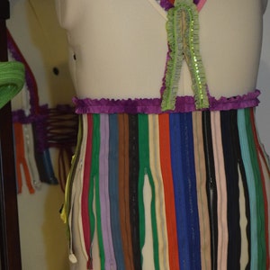 May include: A white dress form with a purple and green top and a skirt made of colorful zippers. The top has a ruffled trim and the skirt is made of many different colors of zippers.
