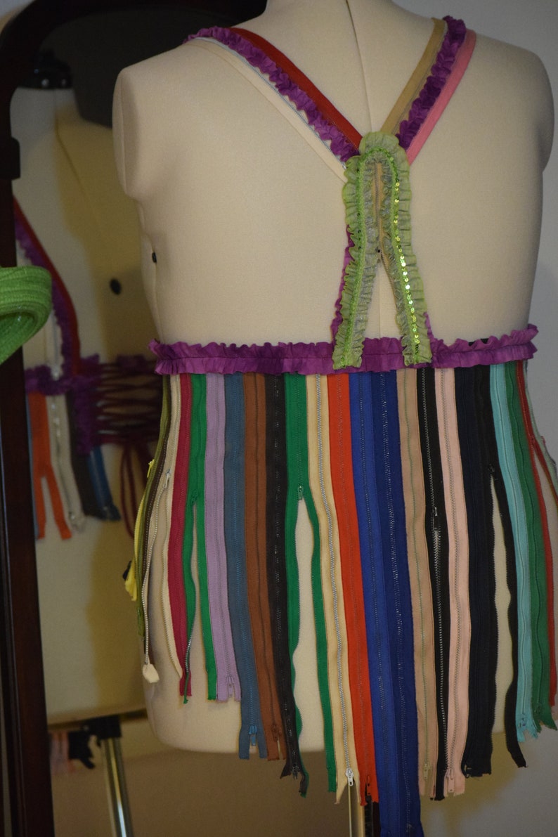 May include: A white dress form with a purple and green top and a skirt made of colorful zippers. The top has a ruffled trim and the skirt is made of many different colors of zippers.