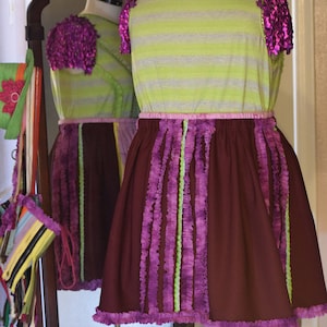 May include: A purple and green striped dress with a brown skirt. The skirt has purple and green ruffles around the bottom and down the sides. The dress has a pink waistband and is on a mannequin.