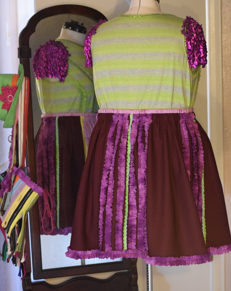 May include: A purple and green striped dress with a brown skirt. The skirt has purple and green ruffles around the bottom and down the sides. The dress has a pink waistband and is on a mannequin.