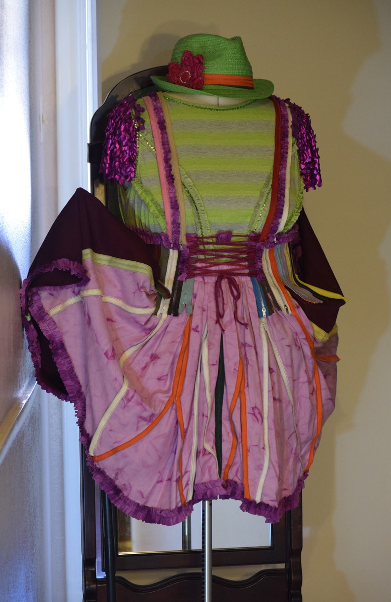 May include: A purple and pink dress with a green hat and a flower on top. The dress is made of different fabrics and has a lot of layers. It is a unique and creative design.