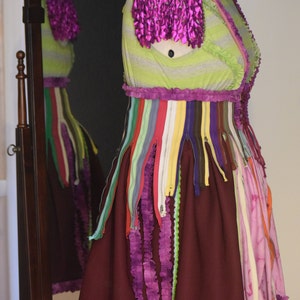 May include: A dress with a green and white striped bodice, a purple ruffle trim, and a burgundy skirt with purple ruffles. The dress is decorated with colorful zippers and is displayed on a mannequin wearing a green hat.