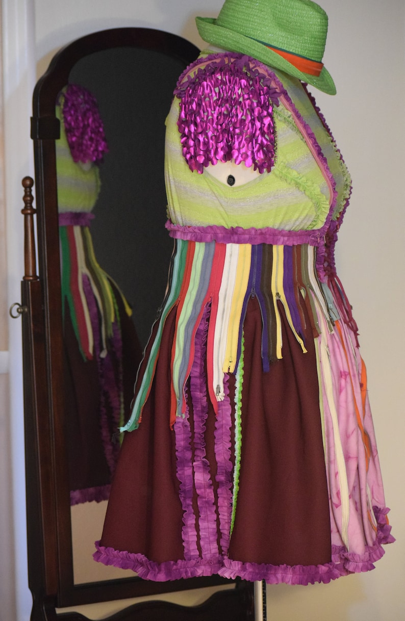 May include: A dress with a green and white striped bodice, a purple ruffle trim, and a burgundy skirt with purple ruffles. The dress is decorated with colorful zippers and is displayed on a mannequin wearing a green hat.