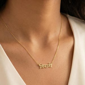 Peut inclure: Un collier doré avec le nom "शिवानी" en écriture hindi. Le collier est porté sur une blouse blanche. Le pendentif est un bijou délicat, suspendu à une fine chaîne.