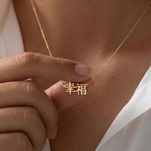 May include: A delicate gold necklace with a pendant featuring Chinese characters. The necklace is held by a person wearing a white button-down shirt. The pendant is in focus, showcasing the intricate design and craftsmanship.