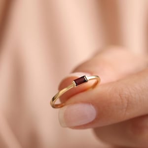 May include: A thin gold ring featuring a single, small, rectangular baguette-cut dark red garnet gemstone set horizontally in the center of the band. The metal band is smooth and polished, showcasing a minimalist design with a deep red stone.