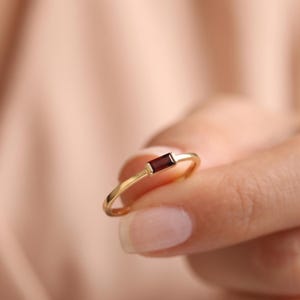 Könnte beinhalten: Ein dünner Goldring mit einem einzelnen, horizontal gefassten, rechteckigen Granat-Edelstein im Baguetteschliff. Die Ringschiene ist glatt und poliert und unterstreicht die tiefrote Farbe des dunkelroten Steins.