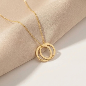 Puede incluir: Un collar dorado con dos círculos entrelazados. Cada círculo lleva grabado un nombre: "Amelia" y "Noah". La fina cadena dorada sostiene el colgante, sobre una tela beige.