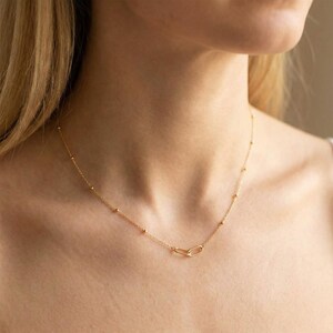 May include: A delicate gold chain necklace featuring small, evenly spaced spherical beads along the length of the chain and a single elongated oval link as a central focal point, resting against the collarbone area of the neck.