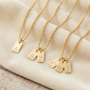 Può includere: Collane color oro con ciondoli rettangolari, ciascuno inciso con una singola lettera. Le collane sono esposte su un tessuto color crema. Le lettere visibili sono A, B, Z, C, H e J.