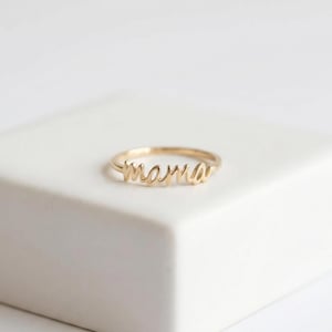 May include: A gold-toned ring featuring the word mama written in a continuous cursive script. The ring has a thin, polished band that connects to the lettering, which forms the focal point of the piece. The design is simple and metallic.