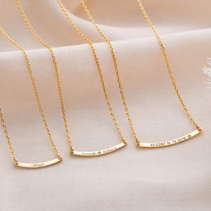 Peut inclure: Colliers dorés avec de fines chaînes et des pendentifs en forme de barre incurvée. Chaque pendentif est gravé d'un nom ou de coordonnées. Les noms "Maggie", "Hannah Rebecca" et les coordonnées "45.0390 N. 79.3079 W" sont visibles.
