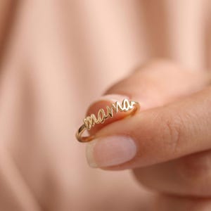 Könnte beinhalten: Ein goldfarbener Ring mit dem Wort mama in geschwungener Schreibschrift. Der Ring hat eine dünne, polierte Ringschiene, und der Schriftzug ist direkt in den oberen Teil des Rings integriert, was ein durchgehendes, fließendes Design ergibt.