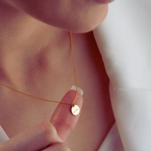 May include: A delicate gold necklace with a small, round pendant. The pendant has the name "Rachel" engraved on it. The necklace is being held up by a finger, and the background is a white shirt.