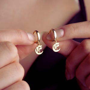 May include: Two gold-coloured hoop earrings, each with a hanging letter "E" pendant in a gothic font. The earrings are displayed against a blurred background, highlighting their polished finish and design.