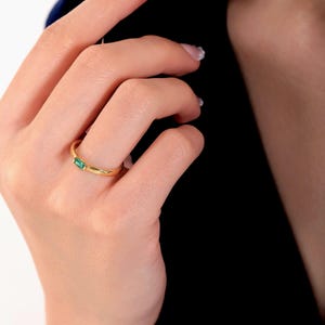 May include: A gold ring with a rectangular emerald gemstone. The ring is simple and elegant, with a thin band and a single, vibrant green stone. The ring is shown on a person's finger.