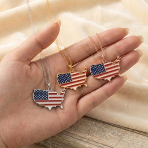 Op de afbeelding: Drie kettingen met hangers in de vorm van de Verenigde Staten, elk met het ontwerp van de Amerikaanse vlag. De hangers zijn in zilver, goud en roségoudtinten, met rode, witte en blauwe emaille. De kettingen worden gepresenteerd op een beige achtergrond.