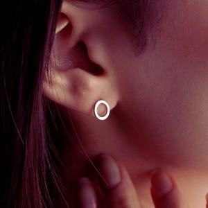 May include: A close-up of a silver open circle stud earring in an ear. The earring is a simple, minimalist design. The earring is made of a shiny metal. The background is a blurred, warm tone.