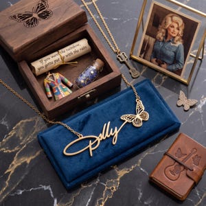 May include: A gold-toned necklace with a script name pendant and a butterfly charm, resting on a blue velvet box. Included are a wooden box with a butterfly lid, a scroll with text, a miniature coat, a glass bottle, a guitar necklace, and a photo.