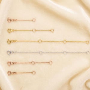 May include: An assortment of jewelry chain extenders in various metallic finishes, including gold, silver, and rose gold. Each extender features a clasp on one end and a small ring on the other, with additional rings along the chain. The extenders are displayed on a cream-colored fabric.