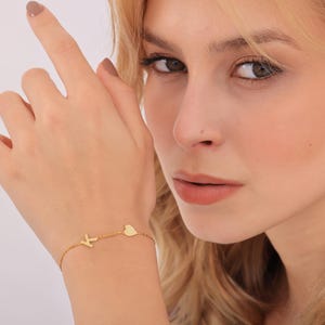 May include: A delicate gold chain bracelet featuring a single capital letter K charm and a small heart-shaped charm. The bracelet is constructed with fine, polished gold links that create a simple and refined look on the wrist, showcasing the letter.