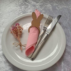 Könnte beinhalten: Ein weißer Teller mit einer rosa Serviette, die von einem hölzernen, hasenförmigen Serviettenring gehalten wird. Eine Gabel und ein Messer befinden sich rechts von der Serviette. Links befindet sich ein kleines Bündel getrockneter Blumen.