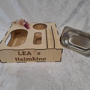 Könnte beinhalten: Eine Snack-Organizer-Box aus hellem Holz mit Aussparungen für Behälter und dem eingravierten Text "LEA's Heimkino" auf der Vorderseite. Eine einzelne herausnehmbare rechteckige Edelstahlschale befindet sich rechts neben der Holzbox.