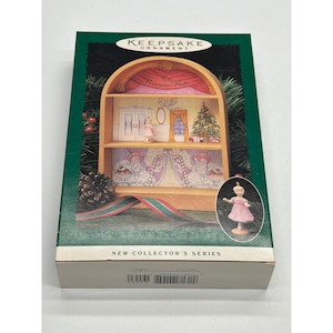 Puede incluir: Una caja verde y blanca con un adorno Keepsake. La caja muestra una estantería de madera en miniatura con un árbol de Navidad, una bailarina y las palabras "The Nutcracker Ballet". También se muestra un pequeño adorno de bailarina.
