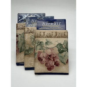 Op de afbeelding: Drie rollen Norwall behangrand verpakt in doorzichtig plastic. Elke rol heeft een decoratieve bovenrand met een beige achtergrond, groene bladeren en trossen paarse druiven. De blauwe kopkaart toont de merknaam Norwall.