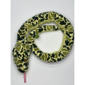 May include: A soft toy snake in green and beige camouflage. The snake has yellow eyes and a pink tongue. A small tag is attached. The toy is designed to look like a snake.