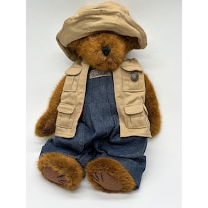 以下が含まれることがあります： ベージュのサファリハット、ポケット付きのベージュのベスト、デニムのオーバーオールを着た茶色のテディベア。ベストには「BEARWEAR」と書かれたパッチが付いています。クマは茶色の毛皮と黒い目をしています。