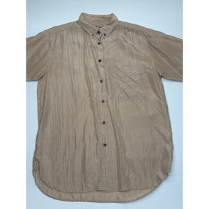 以下が含まれることがあります： ベージュ色のボタンダウンシャツ。襟付きで、胸ポケットが1つ付いています。半袖で、ボタンは濃い色です。生地は少しシワのある質感です。