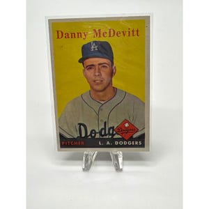 Può includere: Una carta da baseball vintage con Danny McDevitt dei Los Angeles Dodgers. La carta ha uno sfondo giallo con il nome del giocatore in alto. McDevitt indossa un berretto e una maglia dei Dodgers. La carta è in una custodia protettiva.