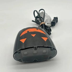 May include: Black Halloween-themed light with orange detailing. The light is shaped like a pumpkin with a carved face design. It has a clear, rounded base with three lights and a black power cord.