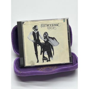 May include: A Fleetwood Mac "Rumours" CD in a clear case, presented in a purple velvet case. The CD cover shows a black and white image of two people. The text "Fleetwood Mac Rumours" is printed on the CD cover.