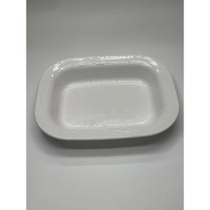 Puede incluir: Una fuente rectangular blanca de cerámica para hornear con esquinas redondeadas. La fuente tiene un borde ligeramente elevado y una superficie lisa y brillante. La fuente está vacía y se encuentra sobre una superficie blanca.