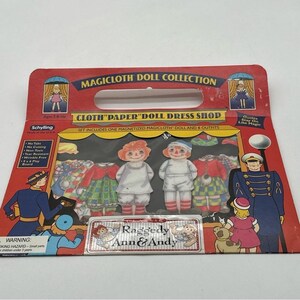 Peut inclure: Un ensemble de collection de poupées Magicloth Raggedy Ann & Andy vintage. L'emballage en carton rouge présente le texte "Cloth Paper Doll Dress Shop" et comprend une poupée aimantée et des tenues. L'ensemble est pour les enfants de 3 ans et plus.