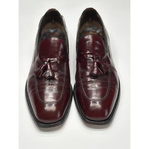 May include: A pair of burgundy leather loafers with a glossy finish. Each shoe features a rounded toe and a decorative tassel. The shoes have a black sole and a low heel. The loafers are displayed on a white background.