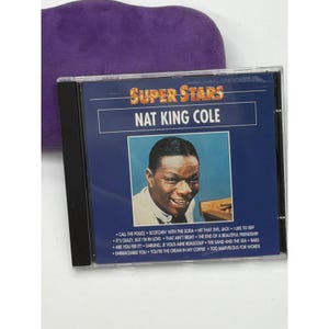 May include: A compact disc case featuring Nat King Cole from the "Super Stars" series. The album cover is a dark blue with a photo of Nat King Cole. Song titles are listed at the bottom. A purple case is in the background.