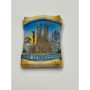 May include: A rectangular souvenir magnet depicting the Barcelona skyline. The magnet shows the Sagrada Familia, other buildings, and the text "Barcelona" against a blue sky, framed by a scroll design.