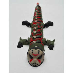 May include: An articulated lizard figurine featuring a beige hockey mask head with red and black details. The body is olive green with red accents and small grey axe motifs along the spine, ending in black limbs and a long, segmented tail structure.