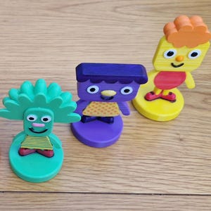 Puede incluir: Tres figuras de juguete de madera de colores con caras sonrientes. Las figuras son verdes, moradas y amarillas, cada una con características únicas. La figura verde tiene una cabeza puntiaguda, la morada tiene una parte superior plana y la amarilla tiene pelo naranja.