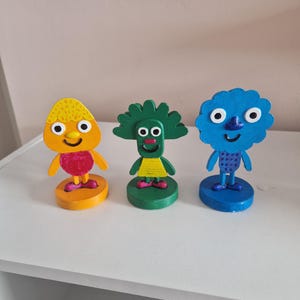 Op de afbeelding: Drie houten poppetjes met beschilderde gezichten en lichamen. Het poppetje links is oranje met een geel hoofd, het middelste poppetje is groen met een gele jurk en het poppetje rechts is blauw met een paars stippenpatroon.