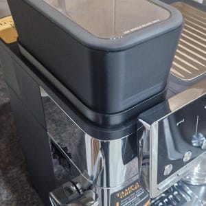 May include: A black and chrome espresso machine with a clear bean hopper. The machine has a digital display and a chrome portafilter. The text "USE WHOLE BEANS ONLY" is visible on the hopper.