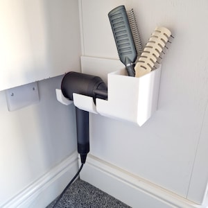 Könnte beinhalten: Weißer, wandmontierter Organizer aus Kunststoff mit einem Haartrockner und drei Haarbürsten. Der Organizer hat zwei Fächer, eines für den Haartrockner und eines für die Bürsten.