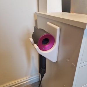 May include: A dark gray and pink hair dryer is mounted on a white wall holder. The hair dryer has a cylindrical body with a pink nozzle and is plugged into a wall outlet. The holder is attached to a white cabinet.