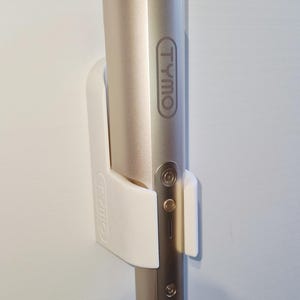 May include: A gold-colored TYMO hair styling tool is mounted on a white holder against a white wall. The tool has a rounded shape with a flower-like design at the top. A gray cord extends from the bottom.