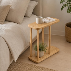 Può includere: Un tavolino in legno di rovere chiaro con piano rettangolare e ripiano inferiore. Una piccola pianta in vaso è appoggiata sul ripiano inferiore. Una tazza e un libro poggiano sulla superficie superiore. Il tavolo è accanto a un letto con biancheria da letto beige.