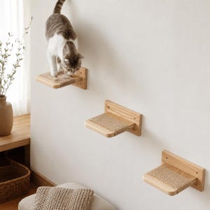 May include: A cat climbs on a set of three wooden wall-mounted cat shelves. Each shelf has a natural fiber scratching surface. The cat is gray and white. The shelves are mounted on a white wall.